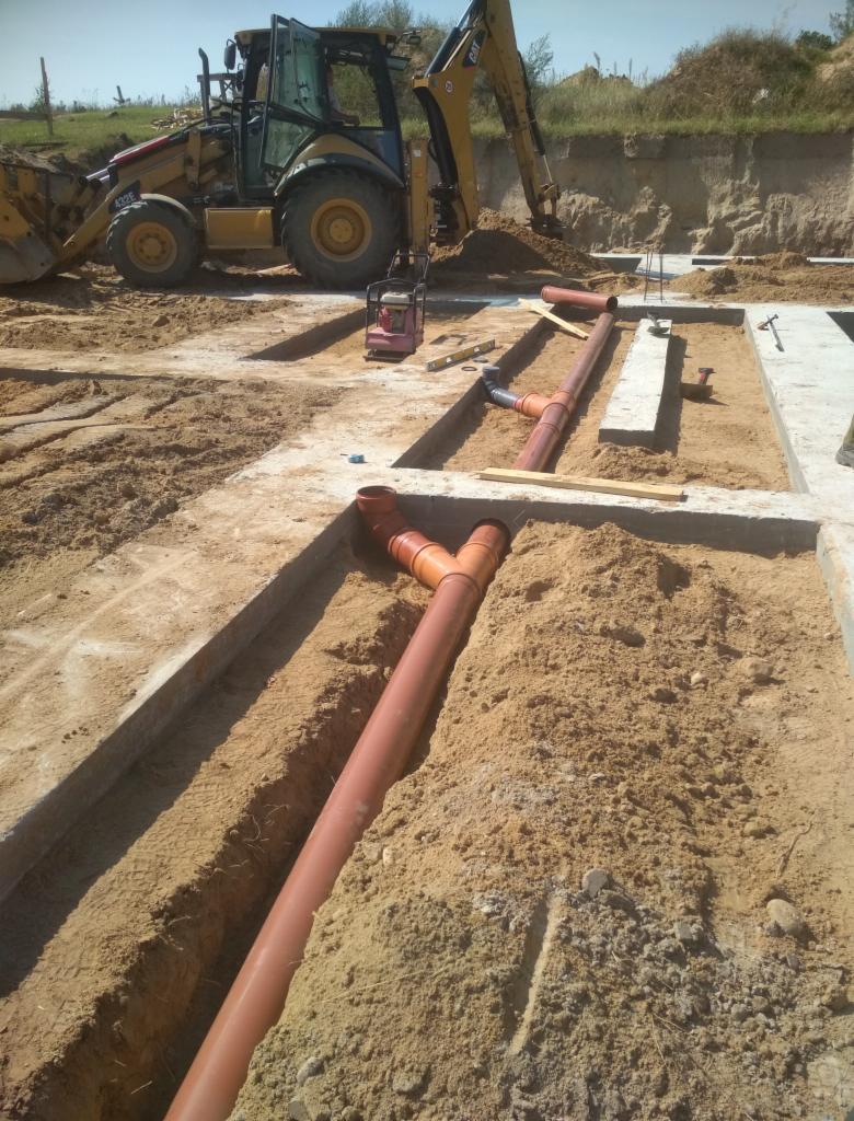Wykop pod fundamenty z ułożonymi rurami kanalizacyjnymi, ubijarką i koparką w tle.