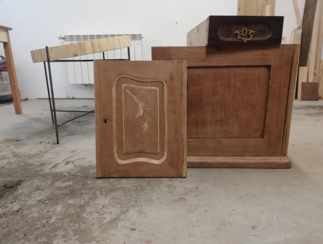 Niedokończona szafka z drewnianymi drzwiczkami zdobionymi motywem roślinnym, obok skrzynia z ozdobnym uchwytem, w tle warsztat.