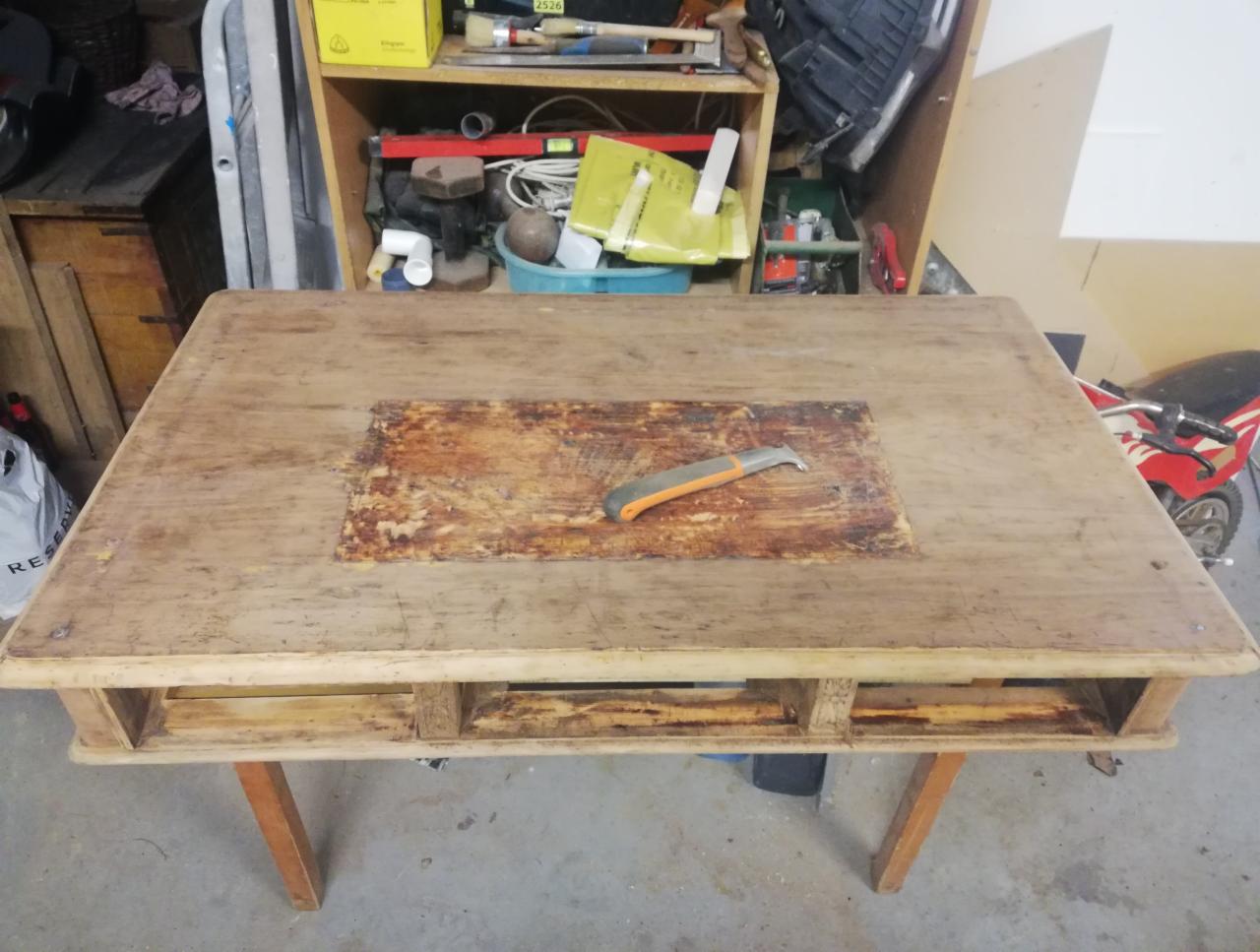 Stary, drewniany stół w trakcie renowacji, z widocznym fragmentem oczyszczonym z farby i narzędziem do usuwania powłok na powierzchni, warsztatowe otoczenie.