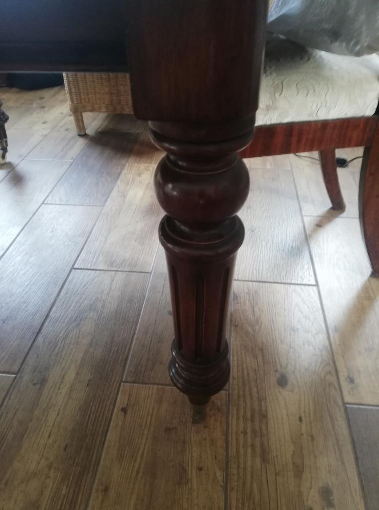 Noga stołu z ciemnego drewna z toczonymi elementami, stojąca na podłodze z paneli imitujących drewno.