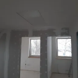 Wnętrze w trakcie prac wykończeniowych: ściany z płyt kartonowo-gipsowych, widoczne łączenia wymagające szpachlowania, sufit z klapą rewizyjną, okna z widokiem na drzewa bez liści.