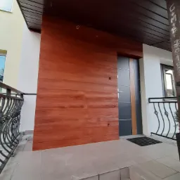 Nowoczesne wejście do domu z elewacją imitującą drewno, ciemnoszara stolarka drzwiowa z brązowymi akcentami i czarną wycieraczką przed drzwiami.