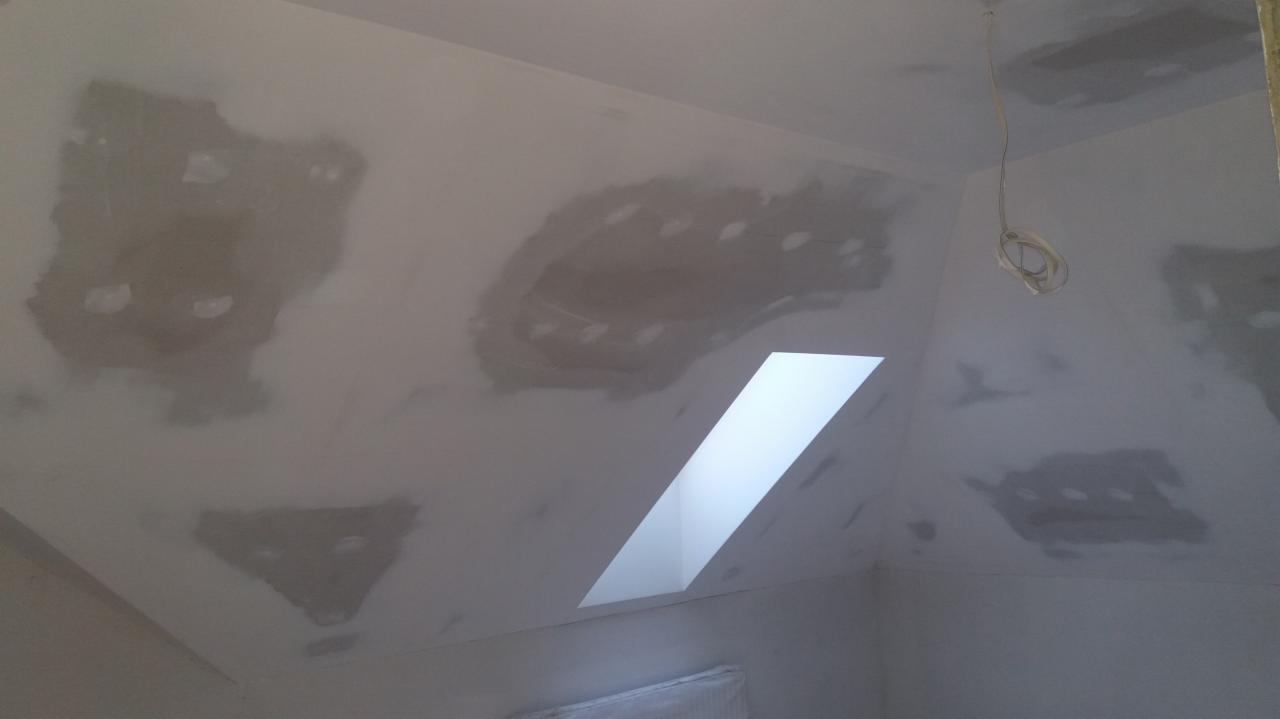 Poddasze w trakcie prac wykończeniowych, widoczne fragmenty płyt kartonowo-gipsowych na skosach i suficie, nierównomiernie pokryte masą szpachlową, z oknem dachowym i zwisającym przewodem...