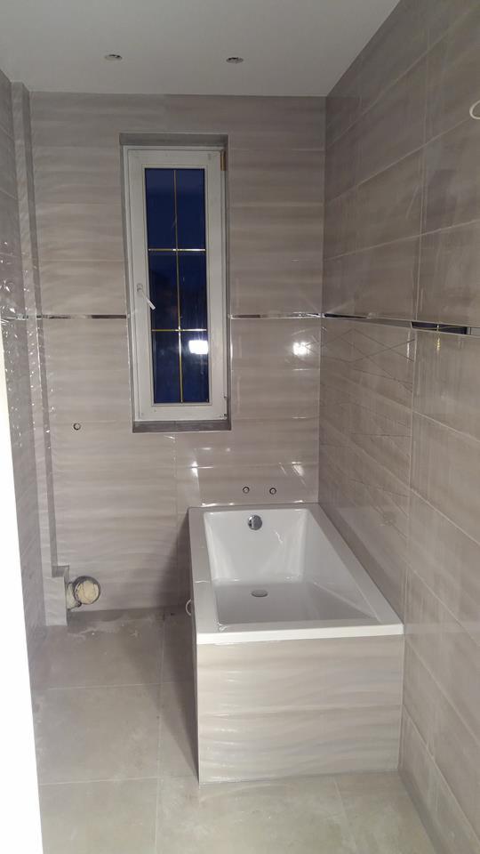 Łazienka w trakcie wykańczania: biała wanna obudowana płytkami imitującymi marmur, ściany wyłożone płytkami w podobnym stylu, małe okno z widokiem na nocne niebo, widoczne niedokończone elementy...