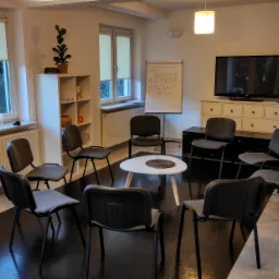 Pokój terapeutyczny z krzesłami ustawionymi w kręgu wokół białego stolika, tablica flipchart z napisem 'Rozżalanie, Użalanie, Po co?' w tle, widok z perspektywy osoby wchodzącej do pomieszczenia.