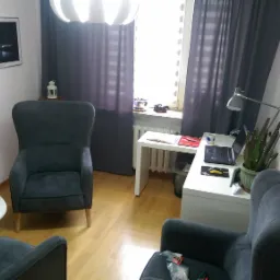 Gabinet z dwoma szarymi fotelami, biurkiem z laptopem, lampką i rośliną, oknem z zasłonami i obrazami na ścianach.