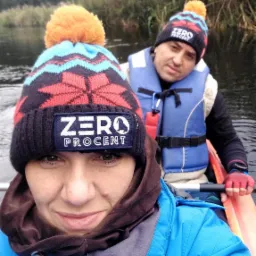 Selfie dw&oacute;ch os&oacute;b w kajaku na rzece, oboje w czapkach z napisem 'ZERO PROCENT' i kamizelkach ratunkowych, zarośla w tle.
