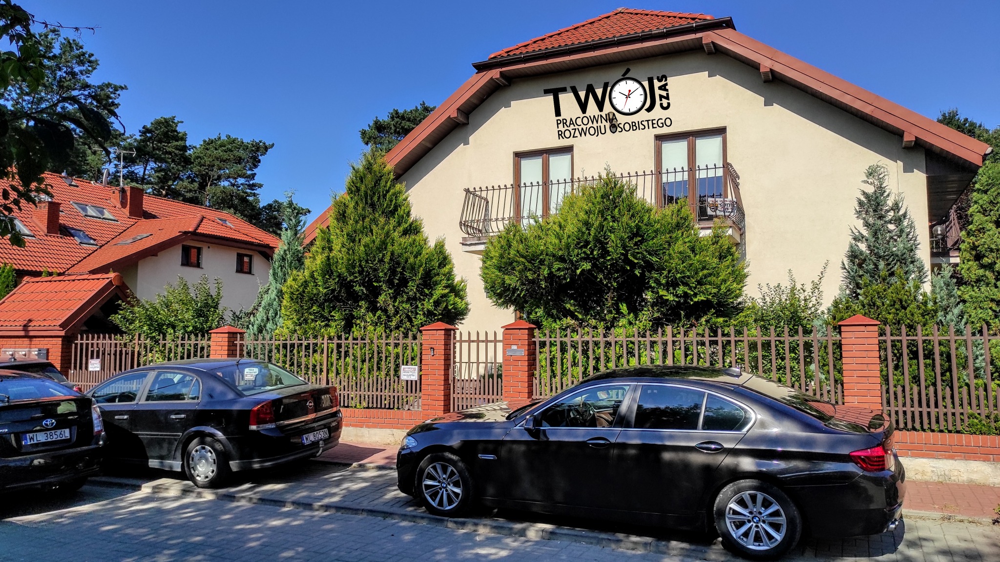 Dwupiętrowy budynek z beżową elewacją i ciemnobrązowym dachem, na ścianie napis 'TWÓJ CZAS Pracownia Rozwoju Osobistego' z symbolem zegara, przed budynkiem zaparkowane dwa czarne samochody...