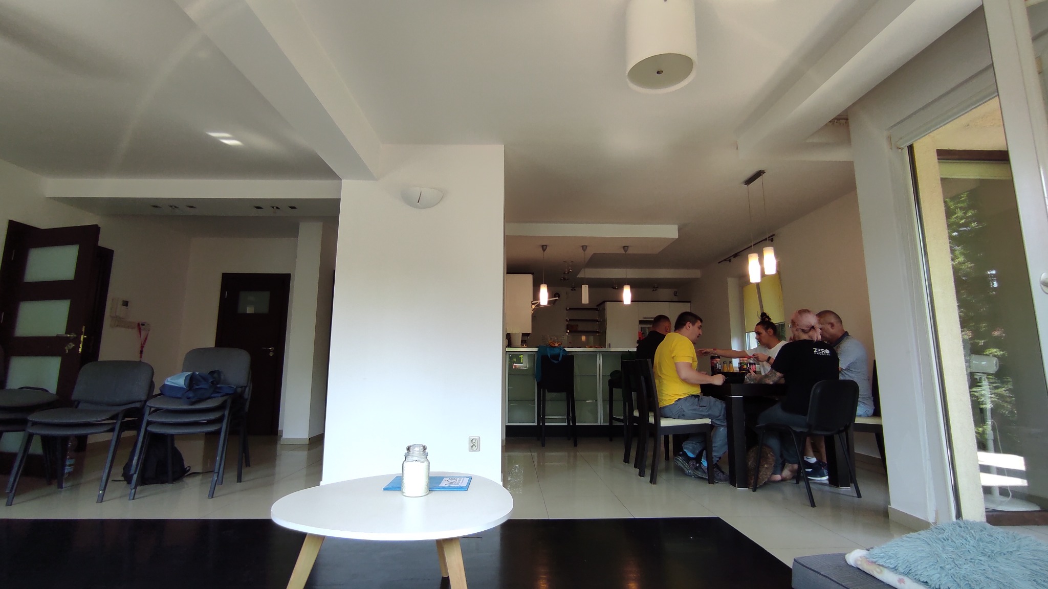 Wnętrze jasnego pomieszczenia z grupą osób siedzących przy stole, prawdopodobnie podczas spotkania lub terapii grupowej, z widoczną kuchnią w tle i słoikiem na małym stoliku z przodu.