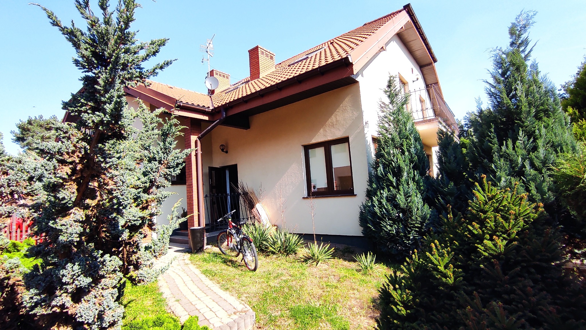 Jasny dom jednorodzinny z beżową elewacją, czerwoną dachówką i balkonem, otoczony iglakami, rower oparty o ścianę przy wejściu.