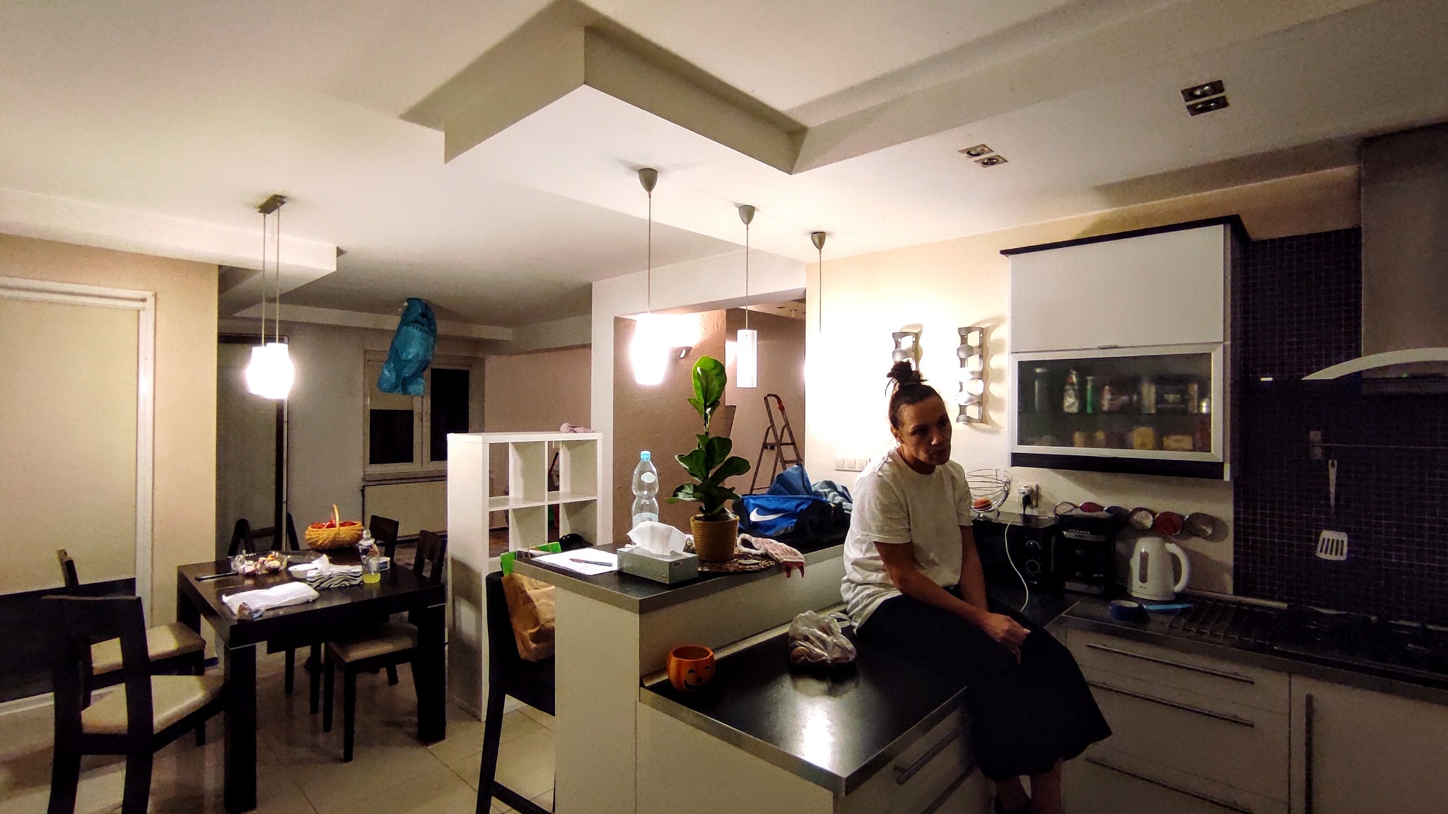 Kobieta siedzi na blacie kuchennym w nieuporządkowanej kuchni z widoczną drabiną, torbą sportową i rośliną doniczkową na wyspie.