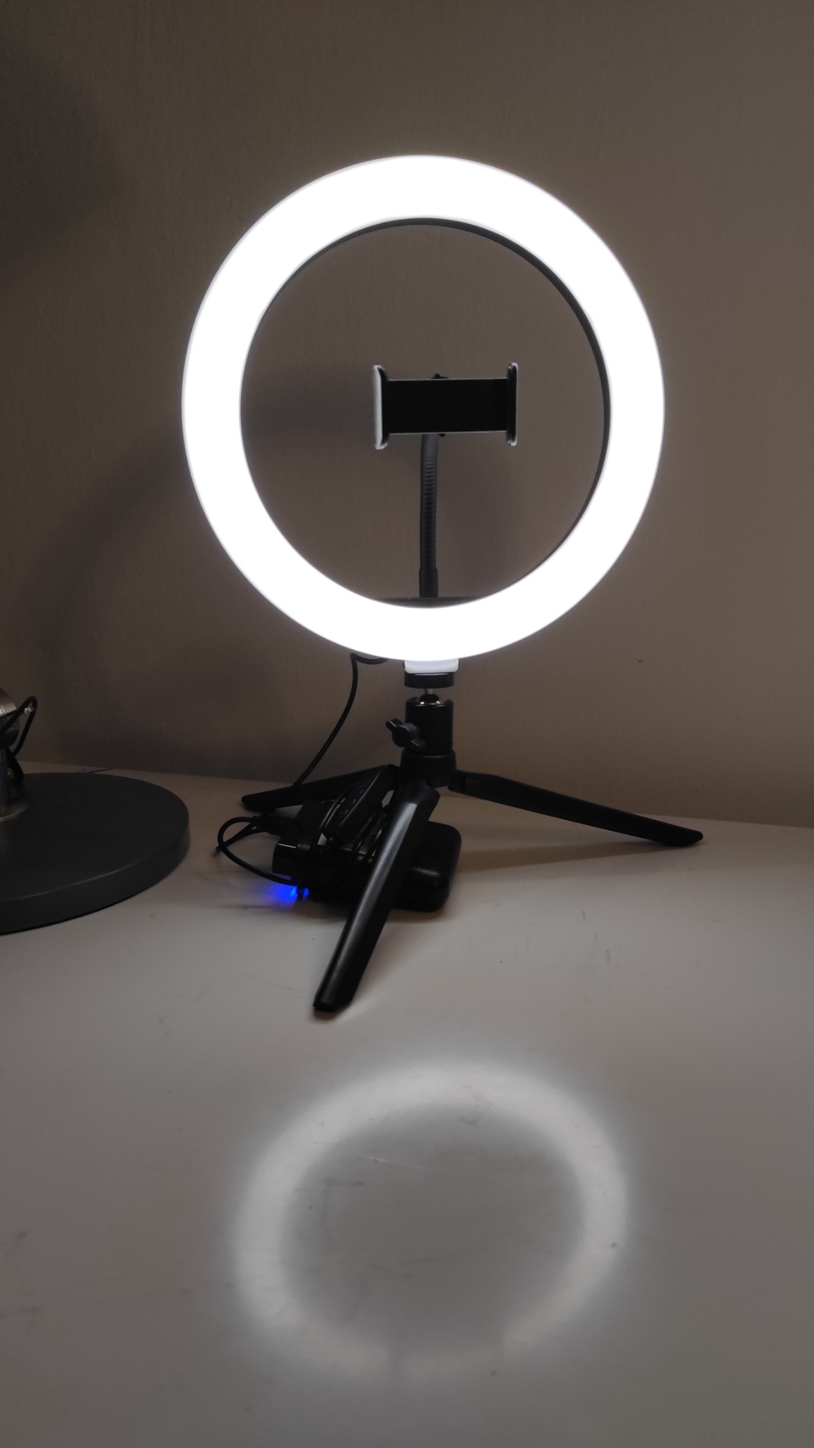 Ring lampa LED na statywie z uchwytem na telefon, z widocznym odbiciem światła na białym blacie.