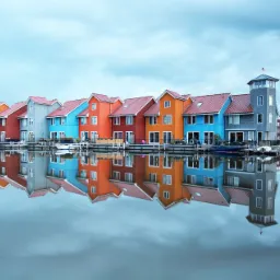 Kolorowe domy na wodzie z czerwonymi dachami, odbijające się w spokojnej tafli jeziora pod pochmurnym niebem.