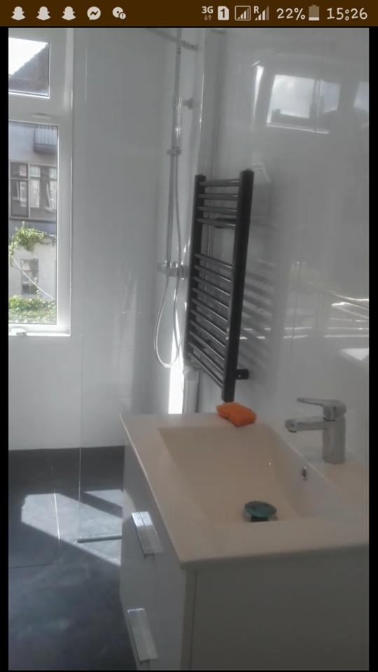 Biała umywalka z szafką, chromowana bateria i czarny grzejnik drabinkowy w wykończonej łazience z jasnymi ścianami i szarą podłogą; widok przez okno na zewnątrz.