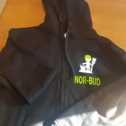 Czarna bluza z kapturem rozpinana na zamek, z zielonym nadrukiem logo firmy NOR-BUD przedstawiającym postać z pędzlem i wiadrem, leżąca na drewnianym stole.