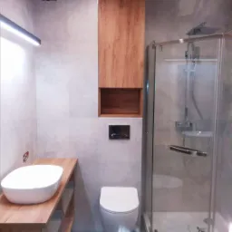 Nowoczesna aranżacja łazienki w Zielonej Górze: szare płytki, drewniane szafki i blat pod umywalką, kabina prysznicowa z chromowanymi elementami, podświetlane lustro.