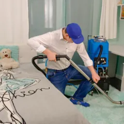 Mężczyzna w niebieskiej czapce i jeansach, używa profesjonalnego urządzenia do czyszczenia jasnoniebieskiego dywanu w pokoju dziecięcym z białymi meblami i pluszowym misiem na łóżku.