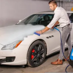 Mężczyzna w szarej koszuli i dżinsach czyści parą biały samochód Maserati, używając niebieskiej ściereczki i niebieskiego urządzenia parowego Optima Steamer w garażu.