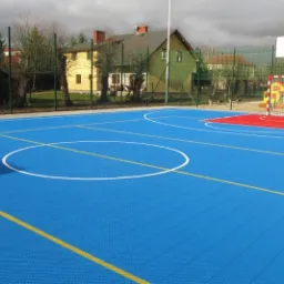 Niebieskie boisko wielofunkcyjne z nawierzchnią modułową, widoczne linie do koszykówki i piłki nożnej, w tle bramka i kosz, całość ogrodzona siatką.