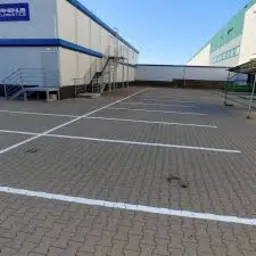 Pusty parking wyłożony szarą kostką brukową z wyraźnie oznaczonymi białymi liniami stanowisk, budynek biurowy w tle, słoneczny dzień.