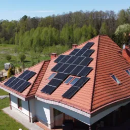 Instalacja paneli słonecznych na dachu domu z czerwoną dachówką, widok z góry na tle zielonego lasu i trawnika, samochód osobowy i dostawczy w tle.