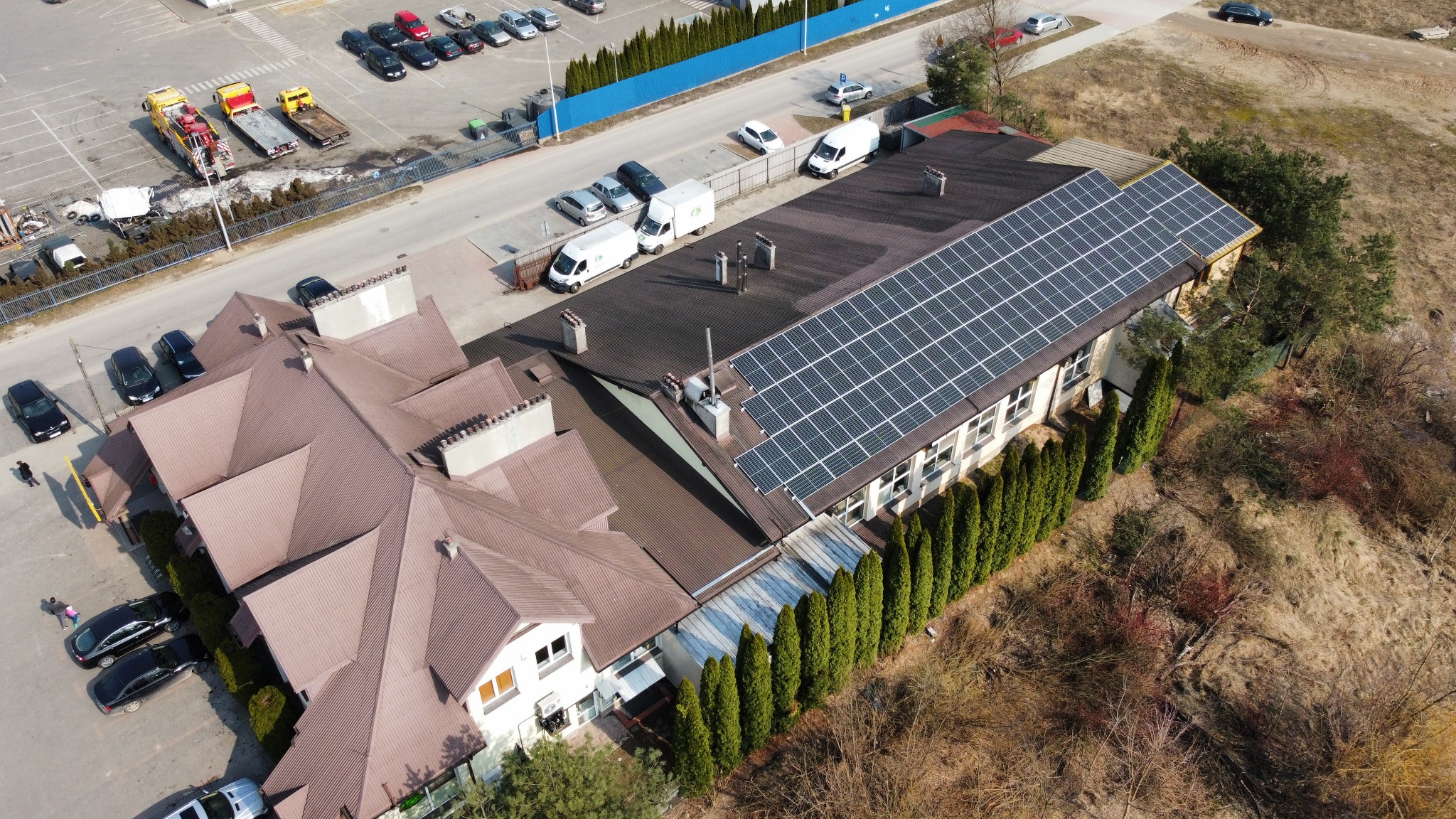 Widok z góry na budynek z panelami słonecznymi na dachu, otoczony tujami, z parkingiem i autolawetami w tle.