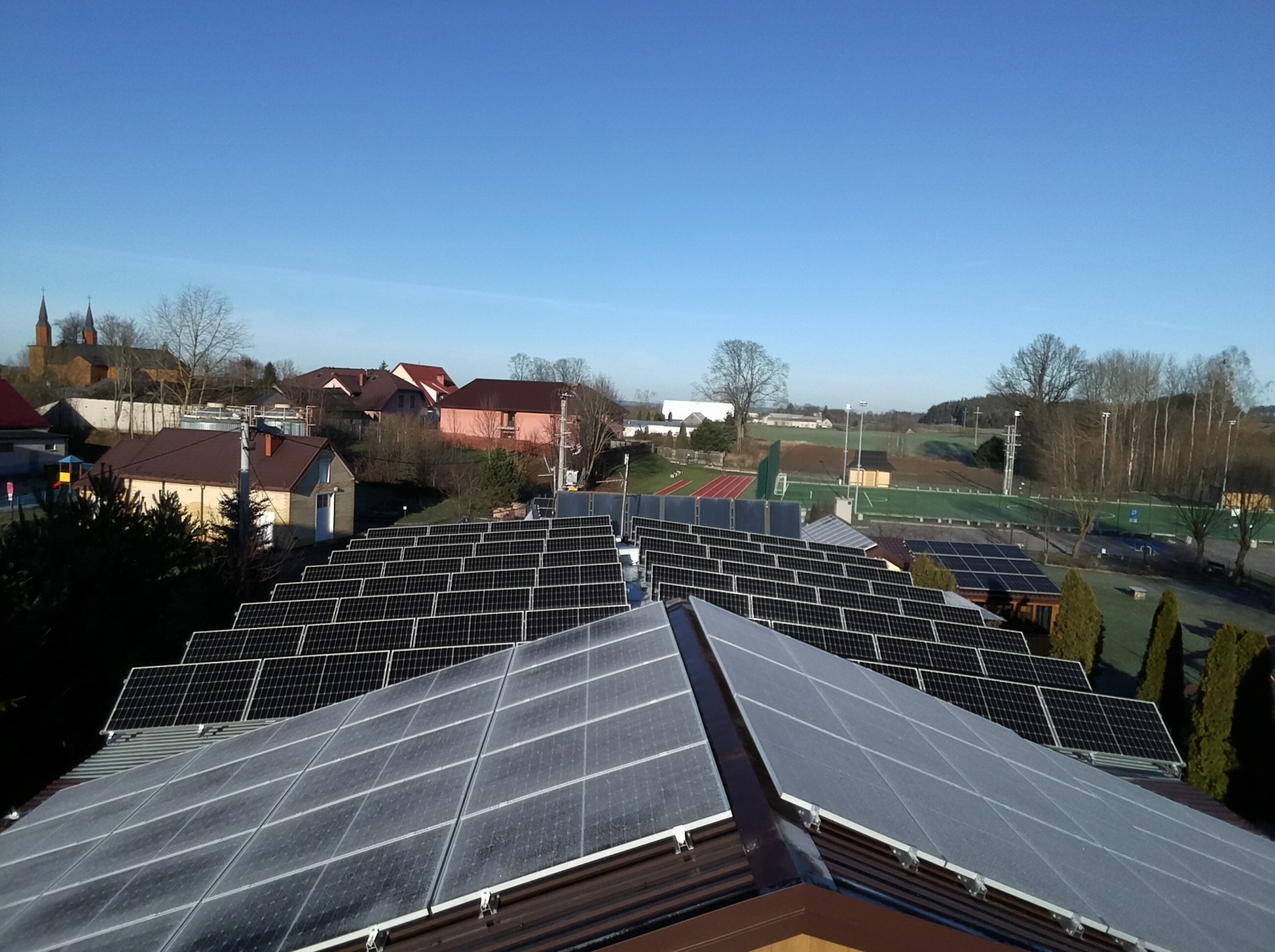 Rozległa instalacja fotowoltaiczna na dachach budynków w wiejskiej scenerii, z widokiem na kościół i boisko sportowe w tle, uchwycona w słoneczny dzień.