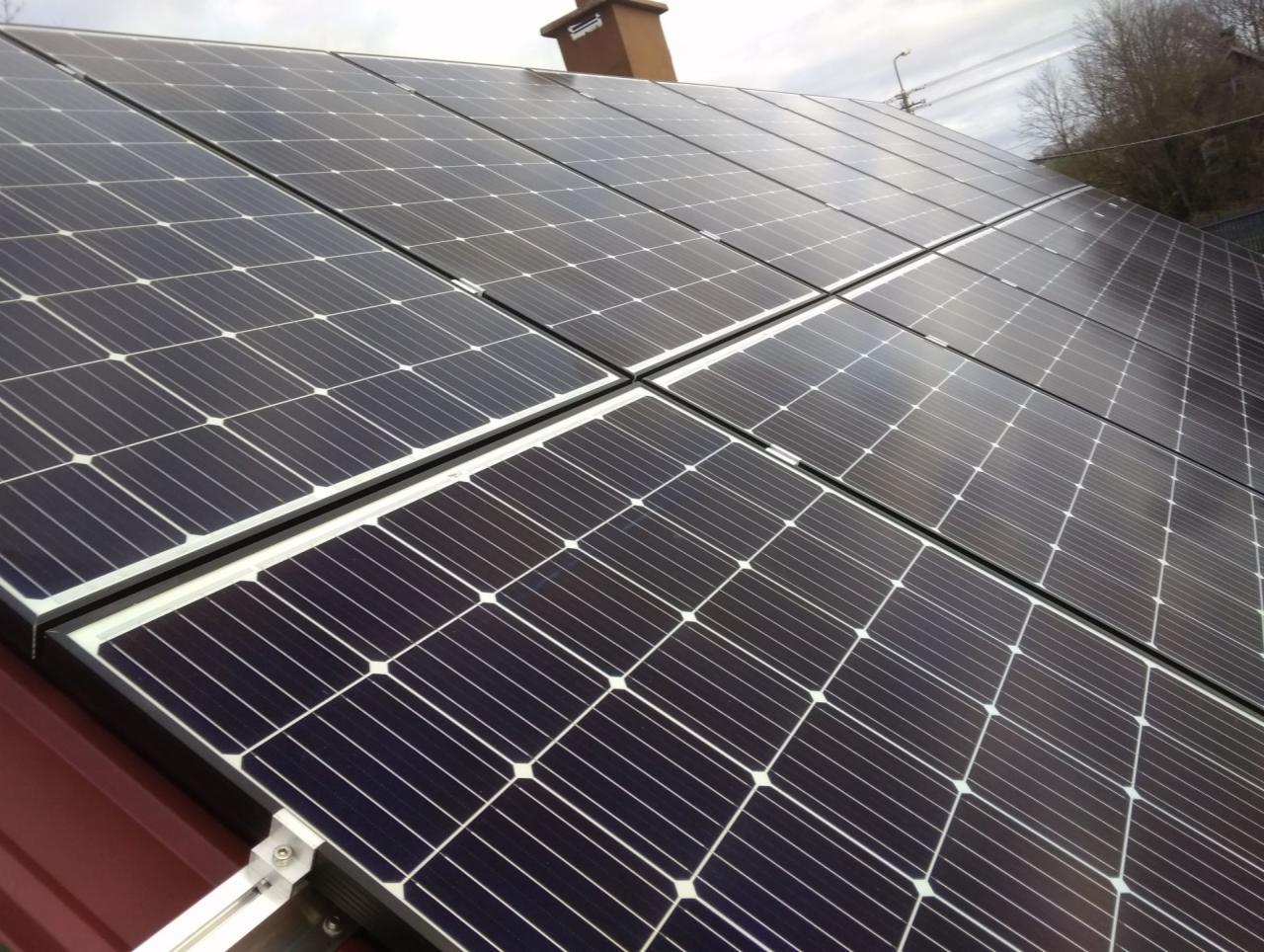 Panele fotowoltaiczne zamontowane na dachu domu z widocznym kominem w tle.