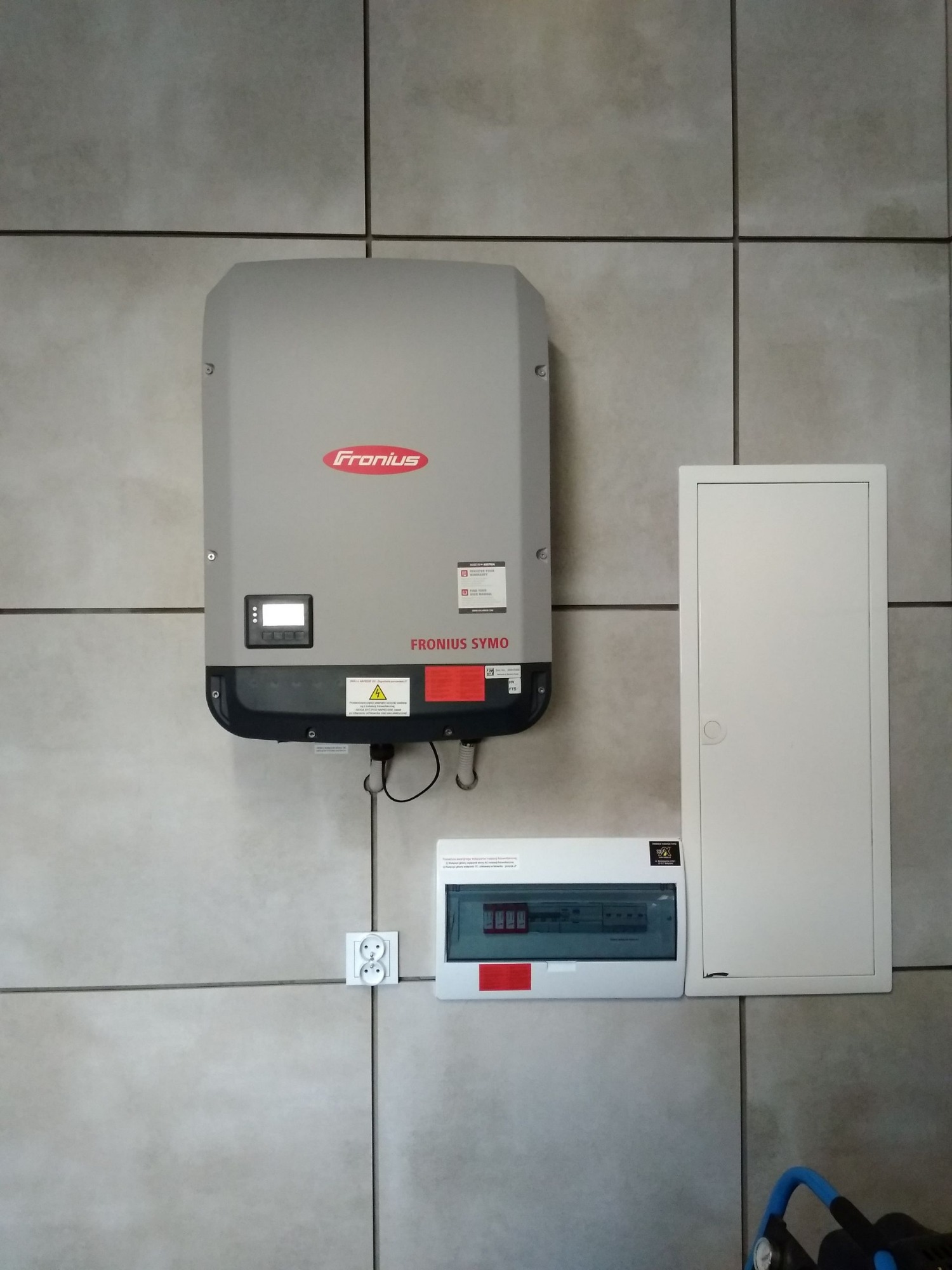 Szary inwerter Fronius Symo zamontowany na kafelkowanej ścianie, obok skrzynka bezpiecznikowa i biała szafka elektryczna.