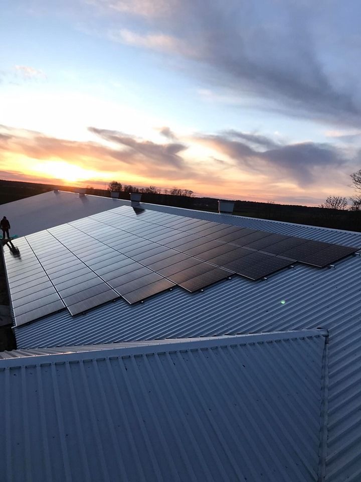 Panel słoneczny na metalowym dachu budynku gospodarczego o zmierzchu, sylwetka osoby w lewym dolnym rogu.