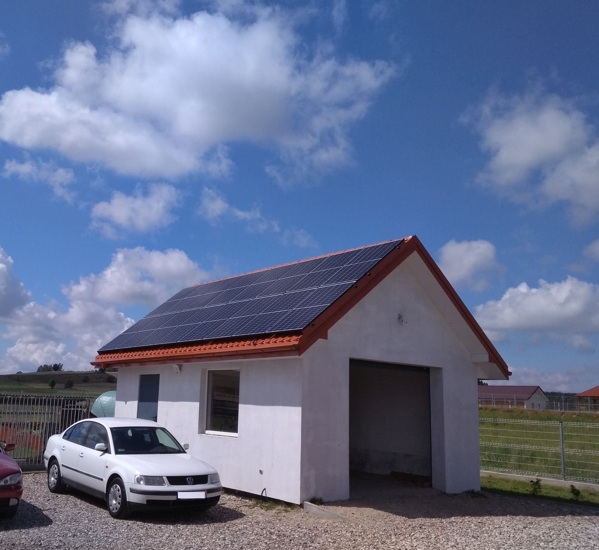 Biały garaż z panelami słonecznymi na dachu, obok zaparkowany biały samochód osobowy, błękitne niebo z chmurami w tle.