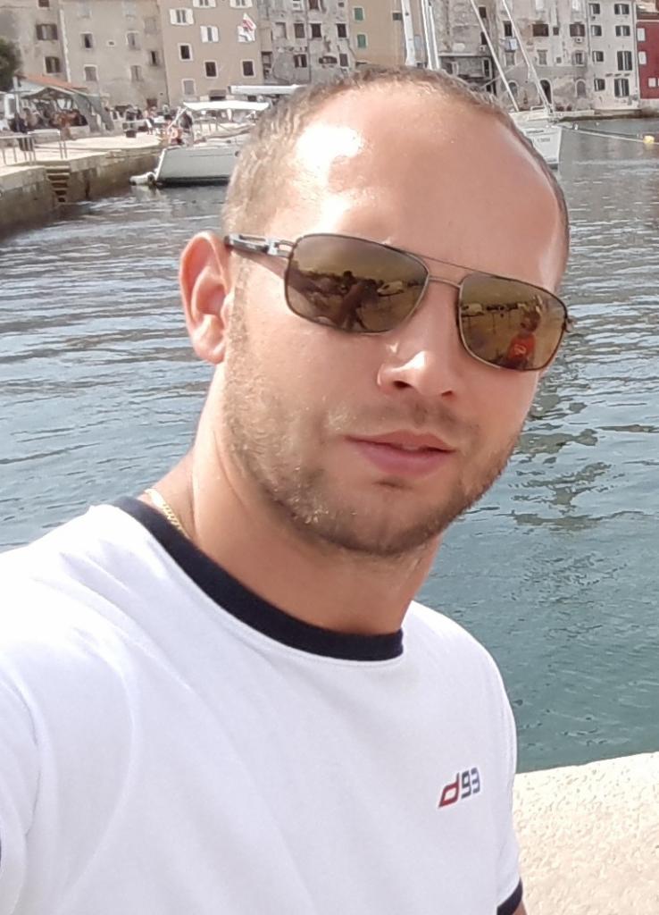 Mężczyzna w okularach przeciwsłonecznych na tle portu z budynkami i łodziami. Odbicia w okularach dodają niepowtarzalnego charakteru.