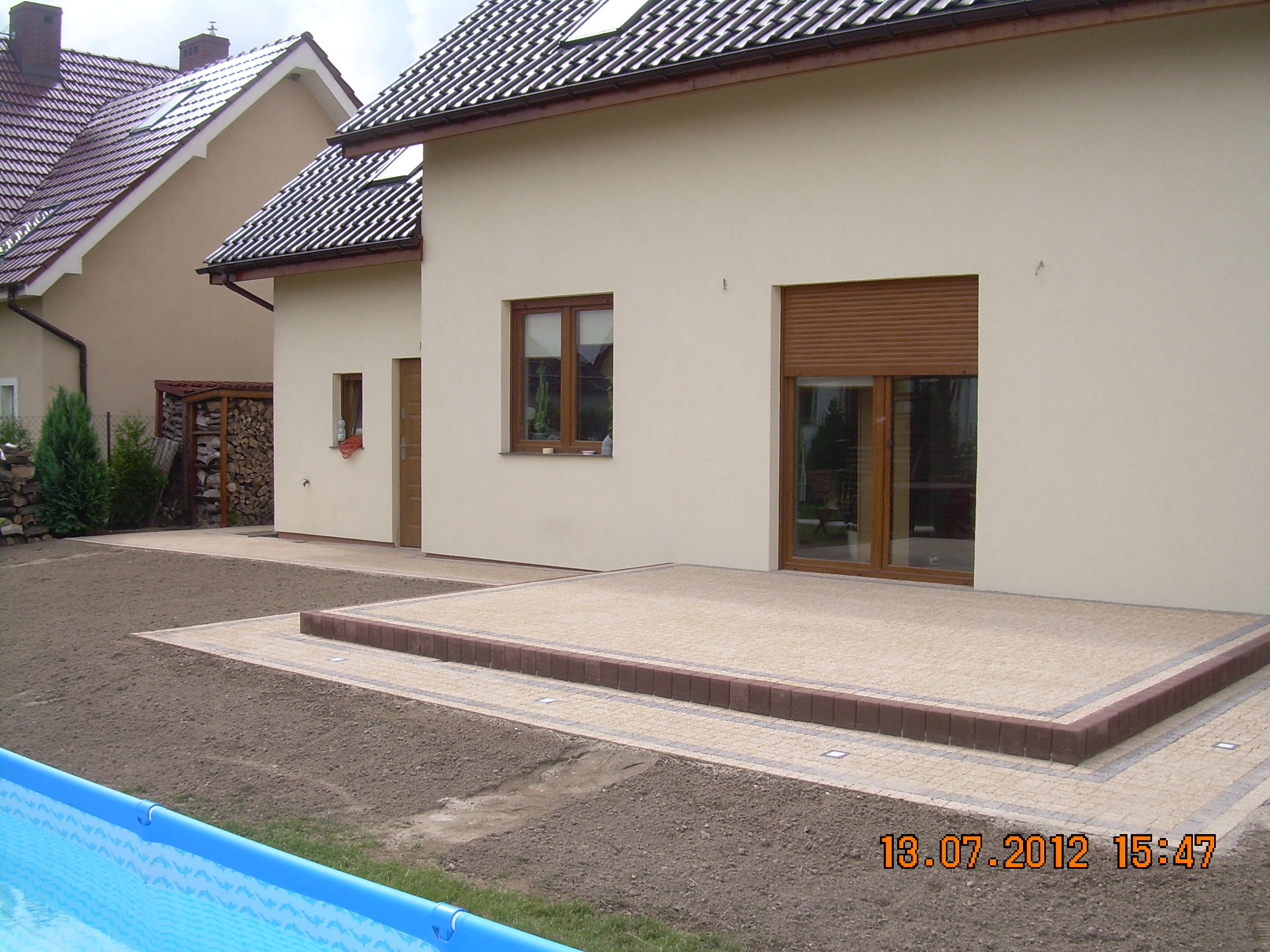 Ułożona kostka brukowa tworząca taras przed domem z brązową obwódką, widoczny fragment niebieskiego basenu ogrodowego, świeżo przygotowany teren pod trawnik.