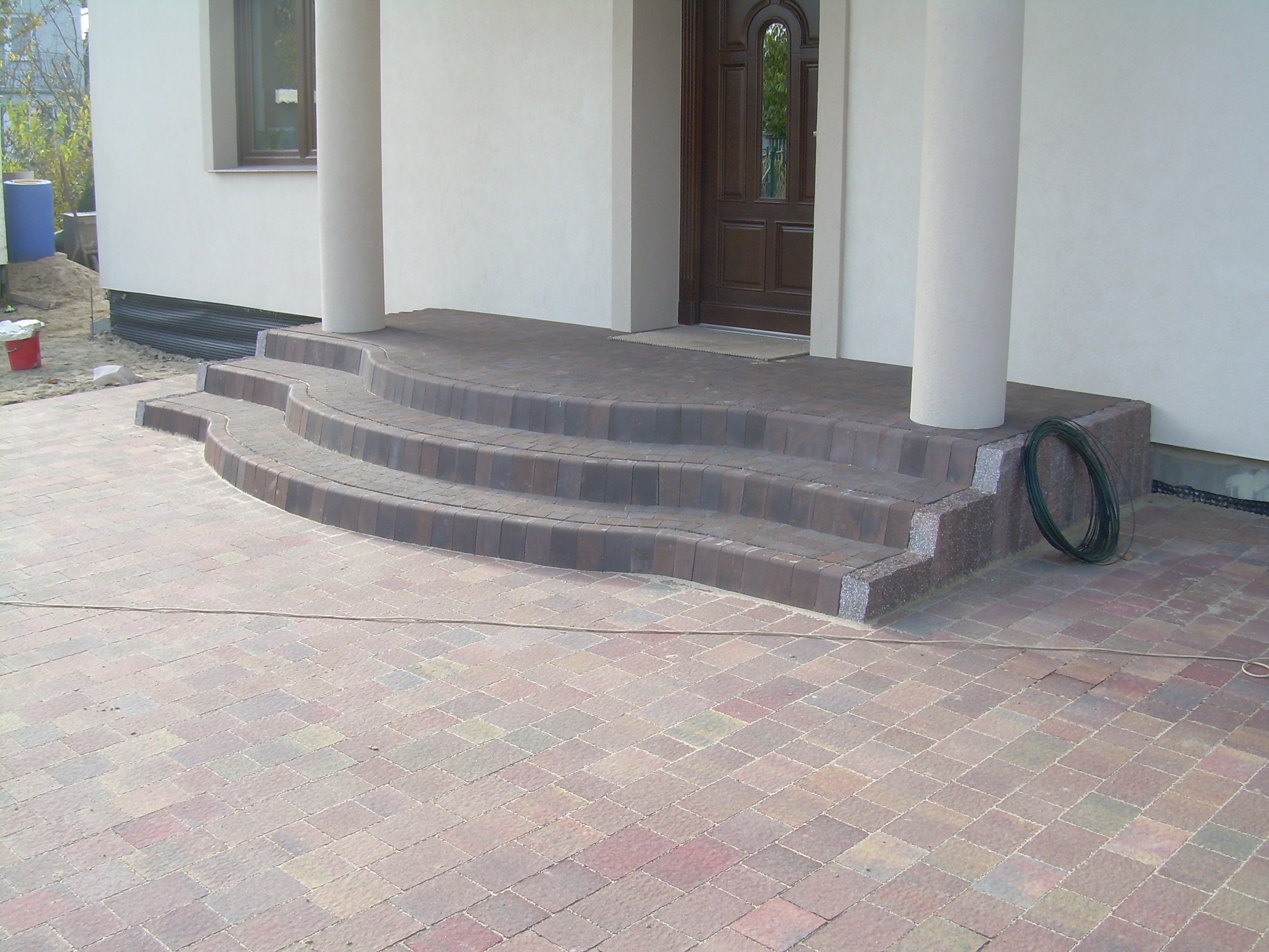 Ułożona kostka brukowa prowadząca do wejścia domu z kolumnami, widoczne trzy półokrągłe stopnie i fragment ułożonego chodnika, obok zwój kabla.