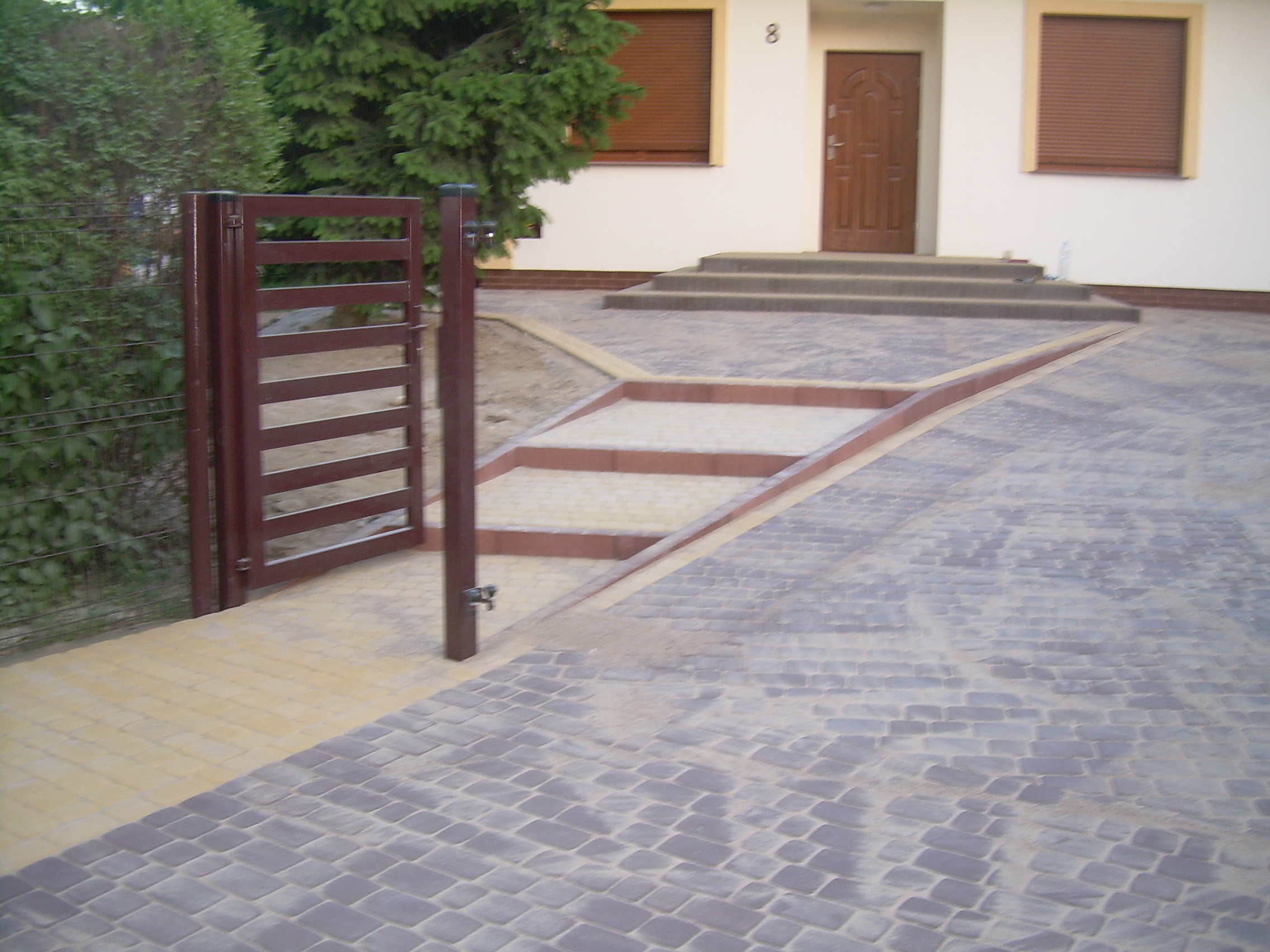 Ułożona szara kostka brukowa prowadząca do domu z brązową bramką i schodami wejściowymi, widok z perspektywy niskiego kąta.