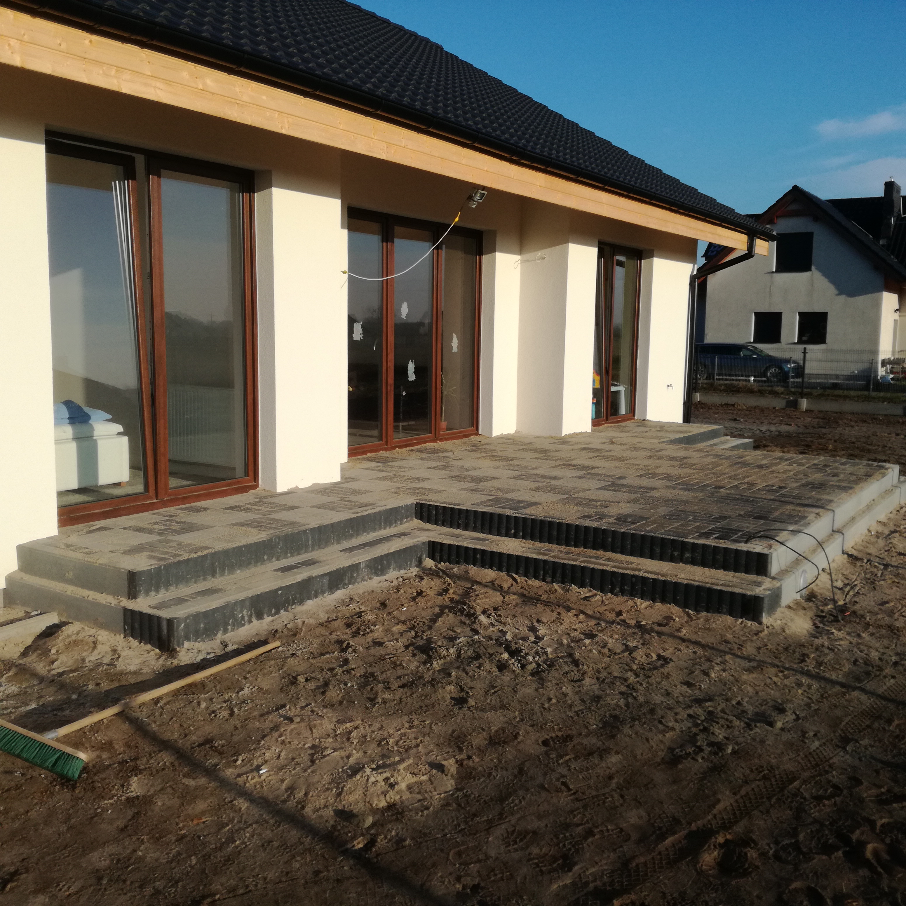 Nowoczesny taras z kostki brukowej w odcieniach szarości, z trzema stopniami, przylegający do jasnego budynku z drewnianymi elementami, w tle widoczny drugi dom i błękitne niebo.