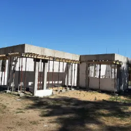 Surowy stan budynku z betonu komórkowego na etapie wznoszenia ścian i szalunków stropowych, widoczne podpory stalowe, teren budowy z piaskiem i trawą, błękitne niebo.