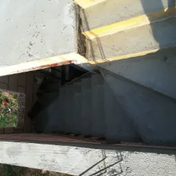 Betonowe schody w trakcie budowy, widok z góry, z widocznym deskowaniem i częściowo wykończonymi stopniami, otoczone ceglaną konstrukcją i zielenią.