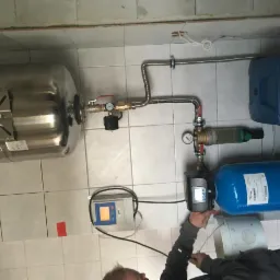 Instalacja systemu uzdatniania wody: zbiornik ciśnieniowy, filtr kolumnowy, panel sterowania i instalacja rurowa na tle wykafelkowanej ściany podczas podłączenia przez instalatora.