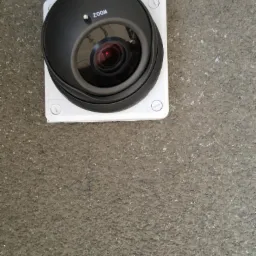 Kopułkowa kamera monitoringu z napisem ZOOM, zamontowana na białej kwadratowej podstawie na szarej, chropowatej ścianie.