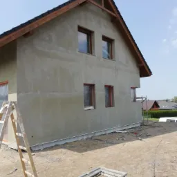 Dom jednorodzinny w trakcie tynkowania elewacji zewnętrznej, widoczne rusztowania i drabina oparta o ścianę, okna zabezpieczone folią, świeżo otynkowana szara ściana.
