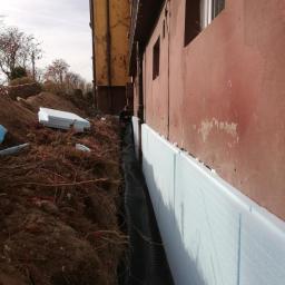 B.R.T. - Odkopane fundamenty budynku z widoczną warstwą izolacji termicznej z jasnoniebieskiego styropianu oraz folią kubełkową, zabezpieczającą przed wilgocią, w tle częściowo widoczny pracownik.