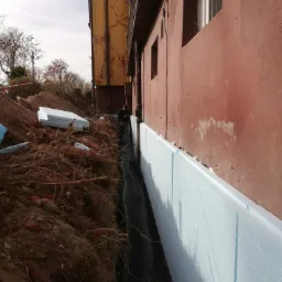 Odkopane fundamenty budynku z widoczną warstwą izolacji termicznej z jasnoniebieskiego styropianu oraz folią kubełkową, zabezpieczającą przed wilgocią, w tle częściowo widoczny pracownik.