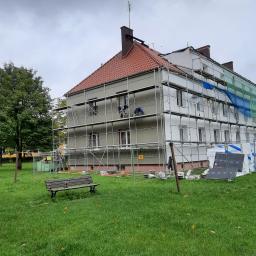 B.R.T. - Pracownik w niebieskim kombinezonie na rusztowaniu, wykonujący prace na elewacji budynku z czerwoną dachówką, widok z trawnika z ławką.