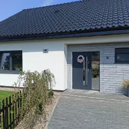 Nowoczesny dom jednorodzinny z ciemną dachówką, białą elewacją i szarą okładziną imitującą cegłę przy wejściu, ozdobiony wieńcem na drzwiach i roślinami w donicach, widok z zewnątrz w słoneczny dzień.