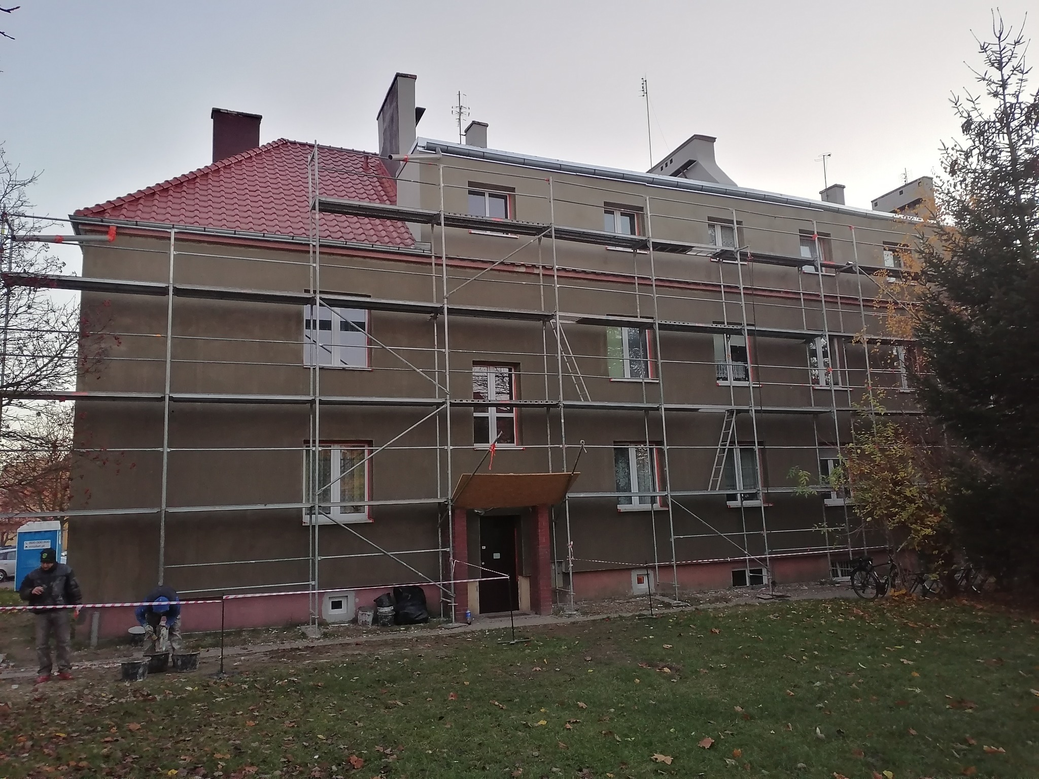 Budynek wielorodzinny w trakcie prac elewacyjnych, z zamontowanym rusztowaniem, widoczne przygotowania do tynkowania i malowania elewacji, pracownicy w trakcie mieszania zaprawy. Częściowo...