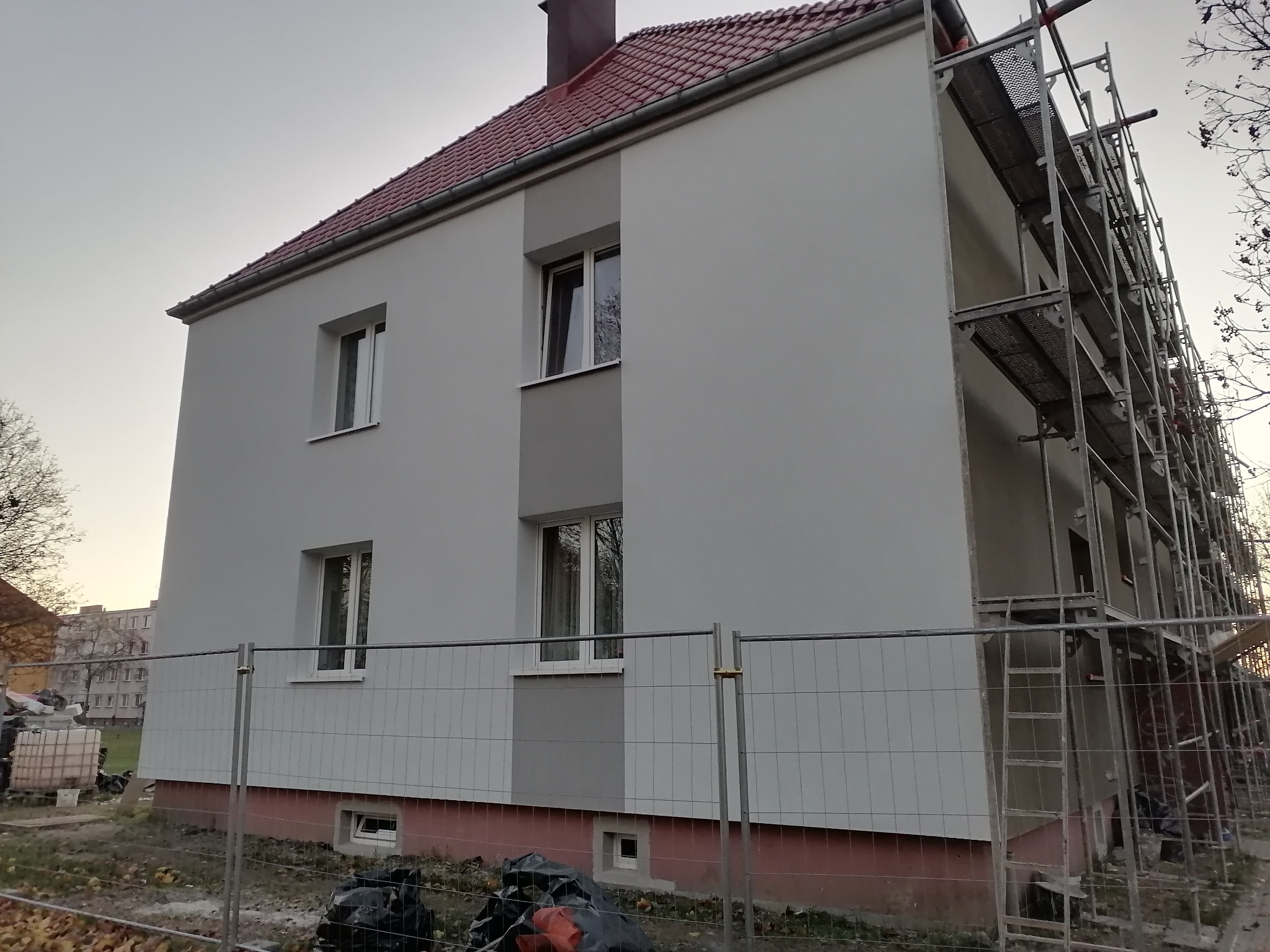 Dwupiętrowy budynek mieszkalny z nową, jasnoszarą elewacją, częściowo osłonięty metalowym ogrodzeniem i rusztowaniem po prawej stronie, z widocznymi workami na śmieci u podstawy.