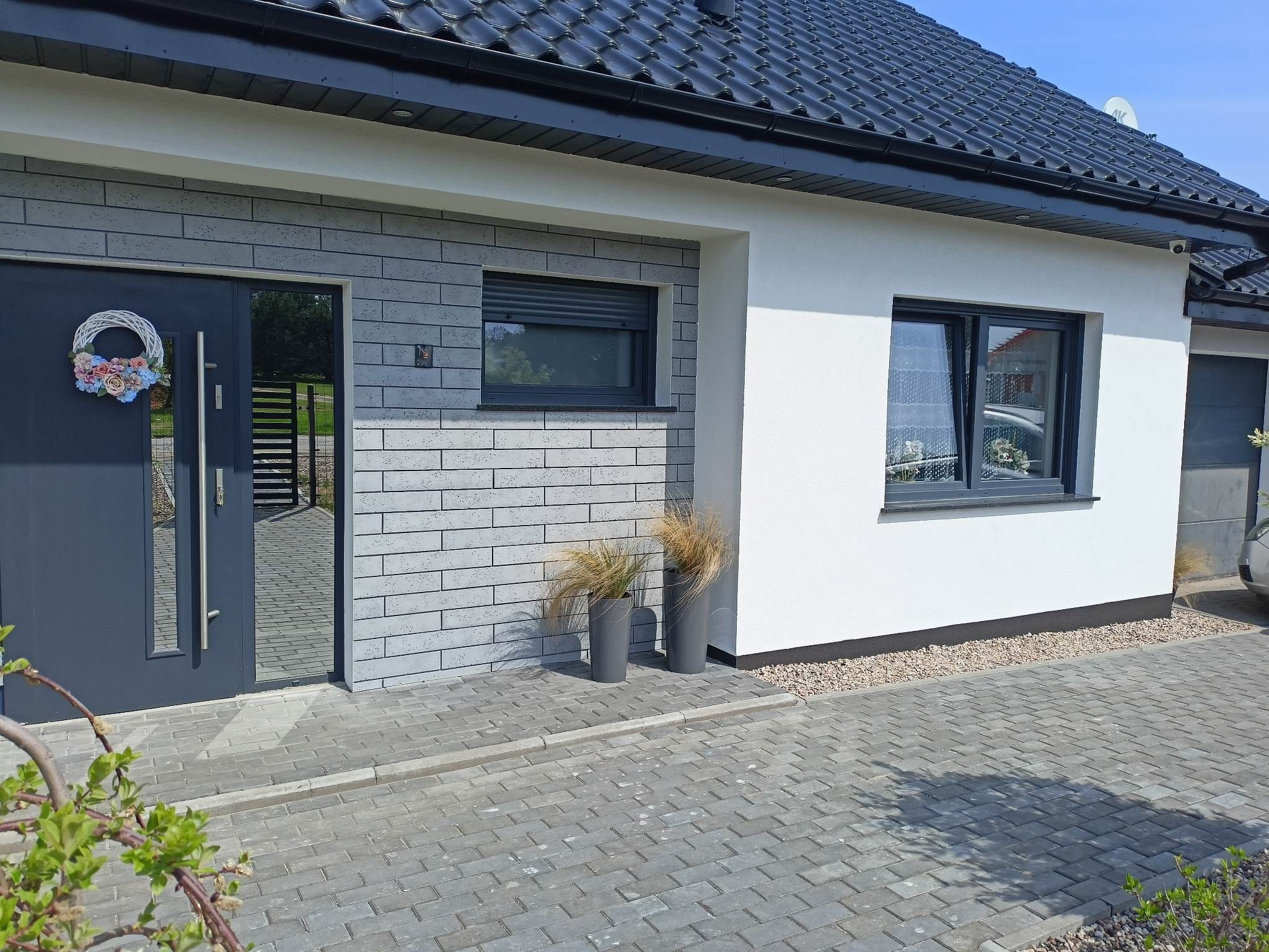 Nowoczesny dom jednorodzinny z elewacją w odcieniach szarości i bieli, ciemna stolarka okienna i drzwiowa, ozdobny wieniec na drzwiach wejściowych, kostka brukowa na podjeździe.