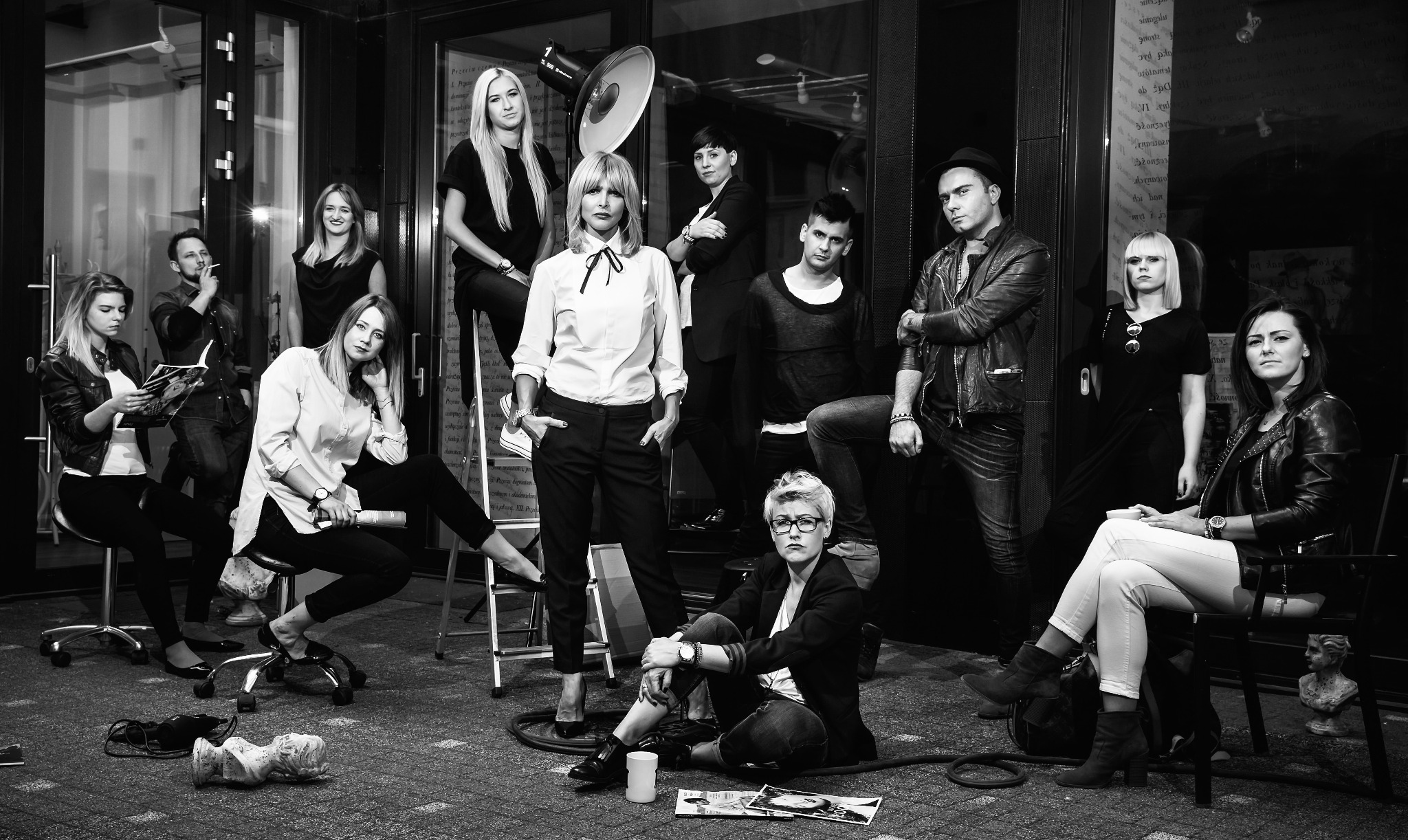 Czarno-białe zdjęcie grupy osób w różnym wieku i ubranych w różnym stylu, pozujących w studiu fotograficznym z lampą błyskową, drabinką i rekwizytami.