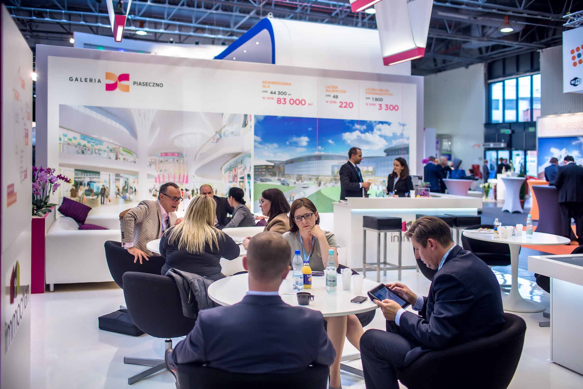 Wnętrze stoiska targowego z osobami siedzącymi przy okrągłych stolikach, na ścianie grafika przedstawiająca wizualizację galerii handlowej Galeria Piaseczno.